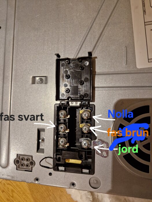 Anslutningsterminal med markerade kablar för nolla, fas brun, jord och fas svart. Grå fas ej inkopplad. Bakgrund av metallisk yta.