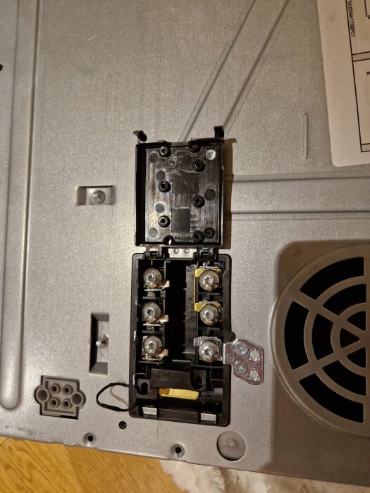 Anslutningsplint för häll med flera skruvterminaler synliga, fastsatta på metallhölje; etikett ovanför och ventilerat område bredvid.