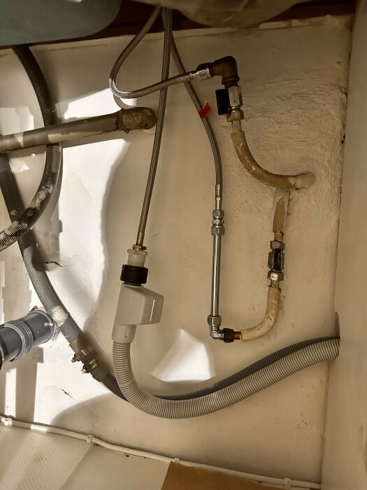 Närbild av köksrör bakom skåp, inklusive vattenledningar och avloppsrör, innan renovering av kök. Installation av nya kök i pågående projekt.