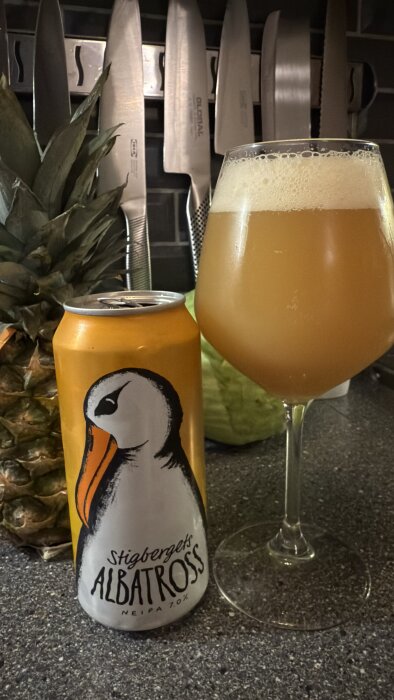 En ölburk med Stigbergets Albatross NEIPA och ett glas med skummande öl på en köksbänk, ananas i bakgrunden.