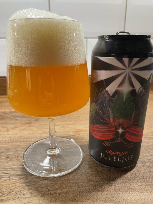 Ett glas skummande julöl bredvid en Stigbergets Juleljus-burk på ett träbord.