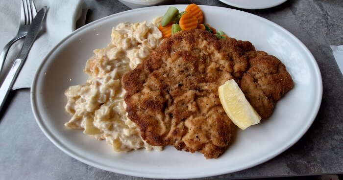 Stor portion schnitzel med potatisgratäng och grönsaker serverad på en tallrik med en citronskiva på toppen.