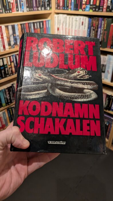 En person håller en bok med titeln "Kodnamn Schakalen" av Robert Ludlum, med en hylla med fler böcker i bakgrunden.