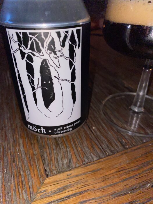 En burk med O/O Brewing mörk robust porter står på ett träbord bredvid ett glas öl.