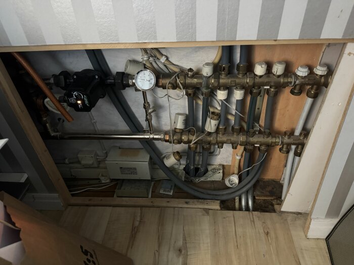 Golvvärmeinstallation med flera rör, ventilkopplingar och en termometer synliga bakom en väggpanel.