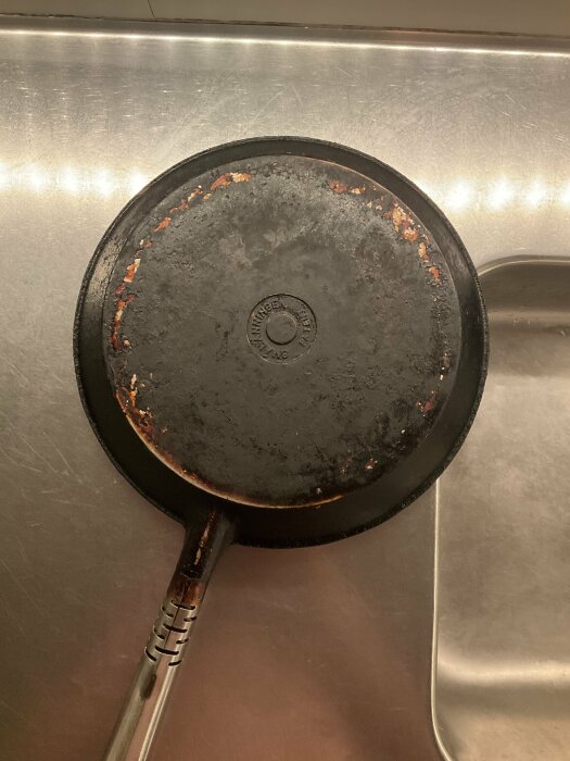 Svart stekpanna med märken på undersidan, placerad på en metallbänk vid en diskho.