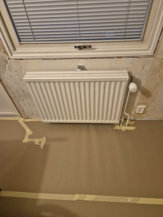 Radiator under fönster, elkontakt på väggen. Tejp markerar området på golvet.