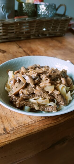 Pasta med entrecote, lök, vitlök, champinjoner och gräddsås i en ljusblå skål på ett träbord.