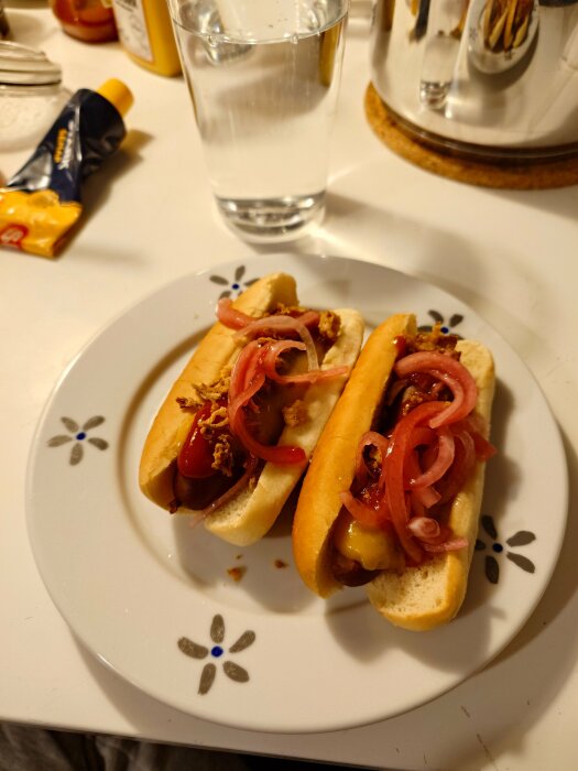 Två korvbröd med senap, ketchup, picklad och rostad lök på tallrik, glas med vatten bredvid.