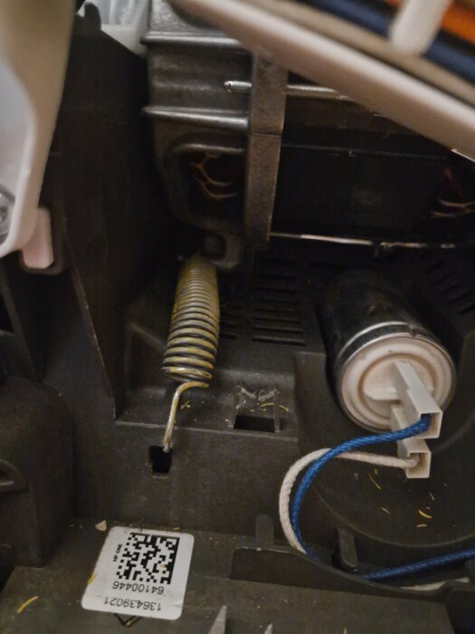 Interiör av mekanisk enhet med en ljusblå och vit kabel, en fjäder och en rem som verkar sitta löst, med en QR-kod synlig i nedre vänstra hörnet.