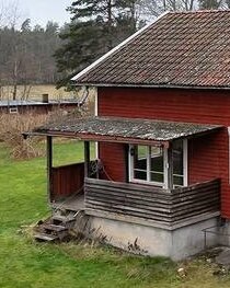 Rött träfärgat hus med tegeltak och en liten altan på en betonggrund, omgiven av grön gräsmatta och med skog i bakgrunden.