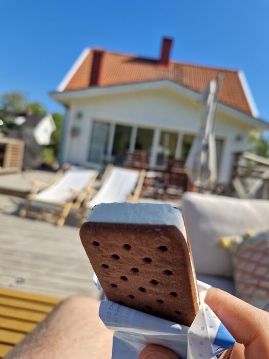 En glasspinne hålls i förgrunden, med utsikt över ett hus med rött tak och veranda i bakgrunden. En solig dag med solstolar ute.