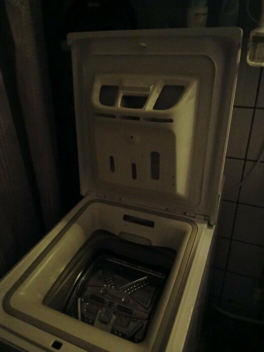 Öppen toppmatad tvättmaskin där doseringsfacket visas, med lock uppfällt, i ett mörkt rum.