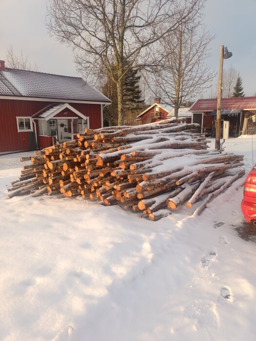 Staplad björkved i snö framför ett rött hus, bredvid ett snötäckt område och ett rött fordon.