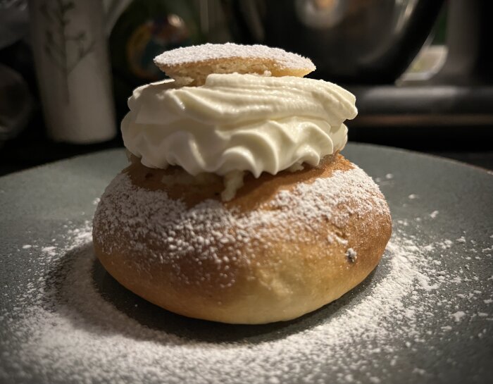 En semla med vispad grädde och lock, pudrad med florsocker, serverad på en tallrik.