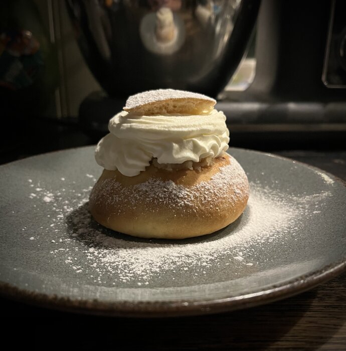Semla på grå tallrik, fylld med grädde och mandelmassa, toppad med florsocker och lock av bröd.