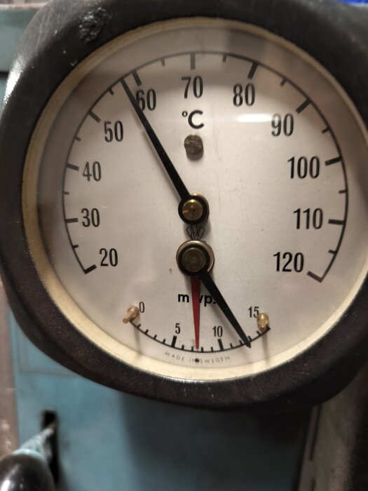 Analog termometer med visare som visar cirka 64 grader Celsius. Tillverkad i Sverige, omgiven av svart ram.