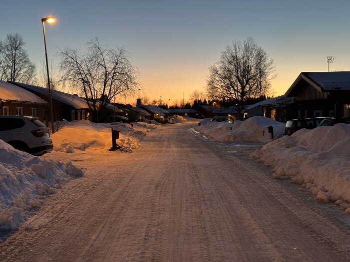 Snötäckt gata vid solnedgång, lyktstolpar lyser upp och hus står längs vägen med träd i bakgrunden. Parkerade bilar syns på sidan av gatan.