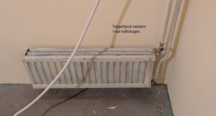 Trippeltjock radiator installerad i en tvättstuga, sett mot en ljusgul vägg med synliga rör och sladdar.