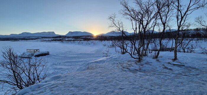 Snötäckt landskap med några träd och ett berg i bakgrunden. Solen är på väg ner vid horisonten.