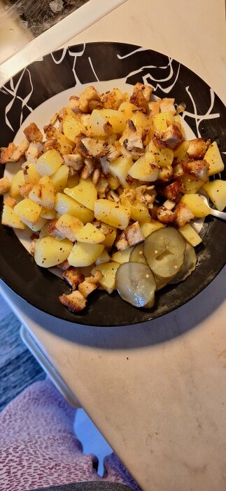Tallrik med pyttipanna gjord på potatis och snitzel, med skivade inlagda gurkor på sidan.