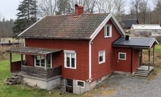 Rött hus med sadeltak och tegelpannor, två våningar. En äldre veranda på baksidan och en liten utbyggnad med plåttak. Skog i bakgrunden.