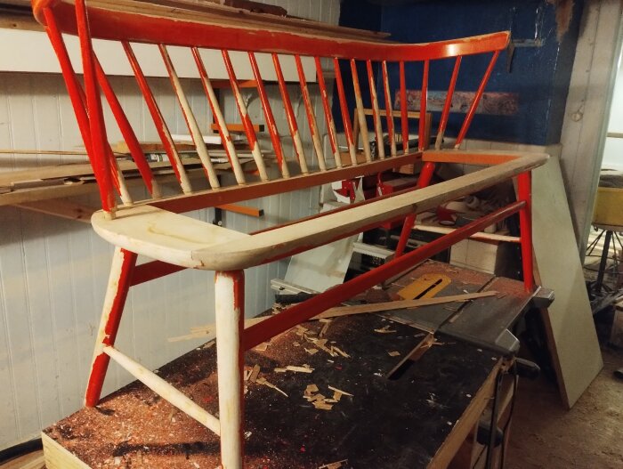Gammal pinnsoffa under renovering, med borttagen plywoodsits och röd plastfärg på träytorna. Verktyg och träflisor på arbetsbord.