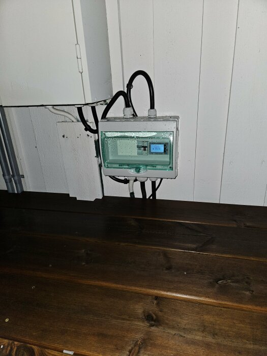 Plastkapsling med mätare monterad lågt på husfasad bredvid mätarskåp, synlig kabeldragning; relaterat till solcellsinstallation.