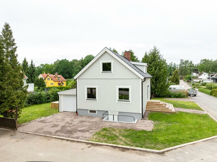 Vit husfasad med garage, nybygge eller renovering, omgivet av träd och gräsmatta, andra villor syns i bakgrunden, i ett bostadsområde.