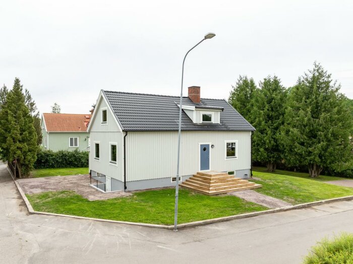 Vit tvåvåningsvilla med grått tak och ny trappa, omgiven av grönska och en gatlykta i en villaområde.