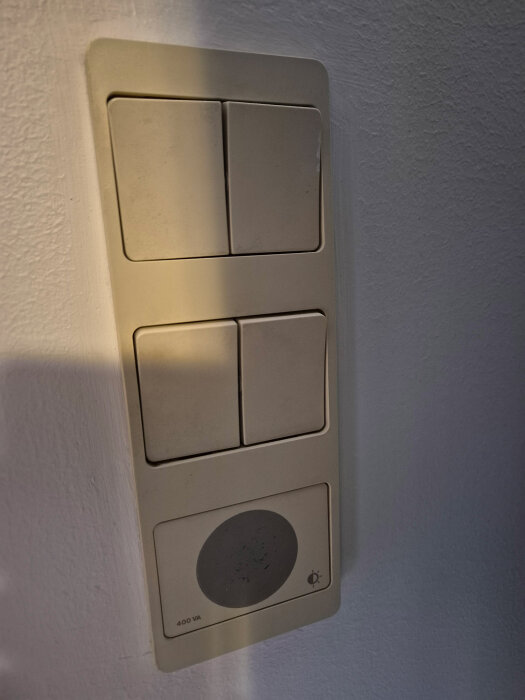 Gammal strömbrytarpanel med fyra vippor och en integrerad rund touch-dimmer, monterad på en ljus vägg.