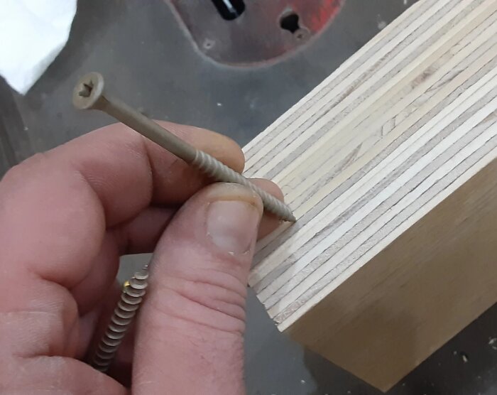 Hand håller en skruv mot kanten av en 18mm plywoodskiva, förbereder för montering av möbelstomme.