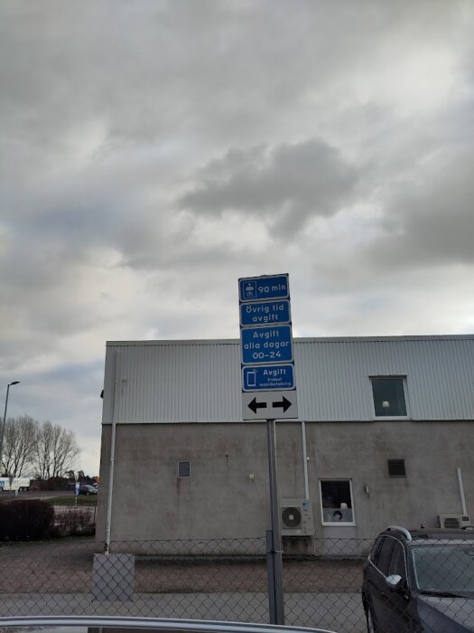 Parkeringsskylt vid industribyggnad med information om tidsbegränsning och avgifter för 90 minuters fri parkering, samt mobilbetalning.