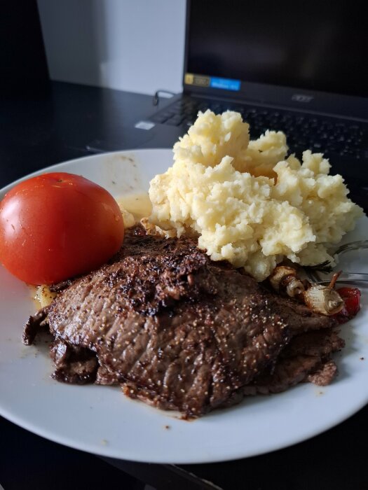 Tallrik med lövbiff, stekt lök, potatismos och tomat, placerad på ett skrivbord framför en laptop.