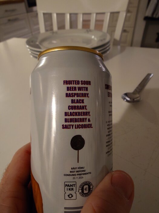En hand håller en burk med text "Fruited sour beer with raspberry, black currant, blackberry, blueberry & salty licorice" i ett kök.
