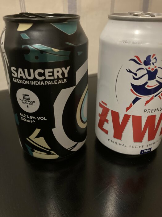 Två ölburkar står på ett bord, en med etiketten "Saucery Session India Pale Ale" och en annan med ett delvis synligt "Żywiec" märke.