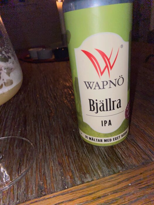 En flaska Bjällra IPA från Wapnö Gårdsbryggeri står på ett träbord, delvis upphällt i ett glas till vänster.