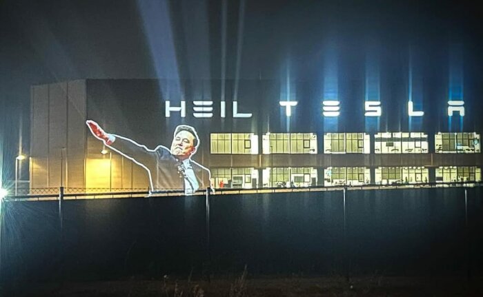 Projektion av en person framför en byggnad med texten "HEIL TESLA" på fasaden, lysande bokstäver i natten.