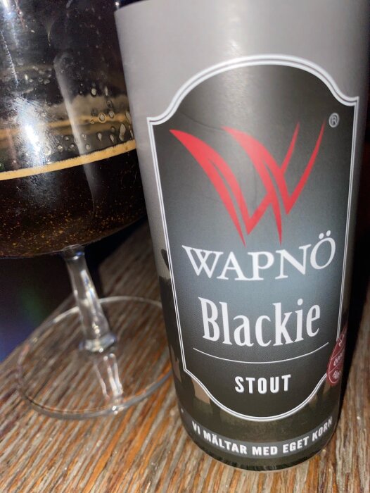 Flaska med Wapnö Blackie Stout och ett glas med mörk öl på ett träbord.