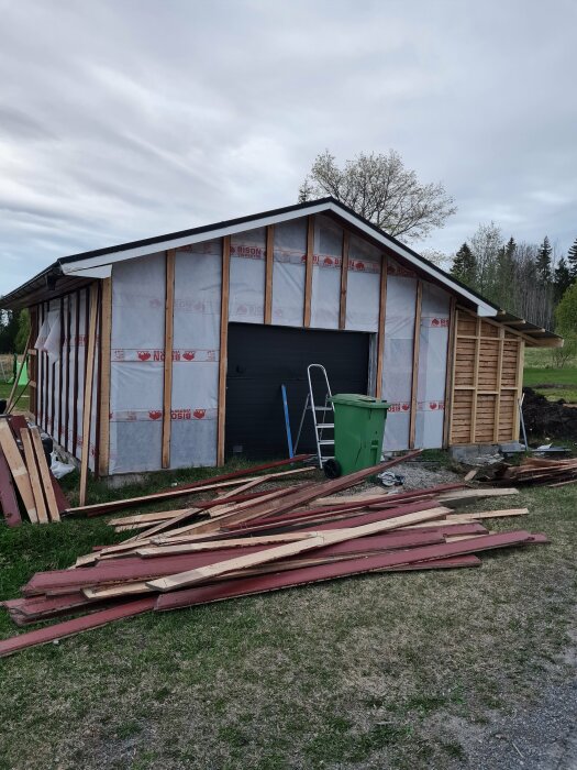 Byggnad under renovering med skyddsplast på väggarna, en grön soptunna, stege och staplat virke bredvid.