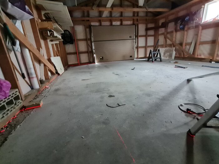 Byggrenovering i garage med betonggolv, träväggar och laserlinjer markerade. Verktyg och material ligger spridda på golvet.