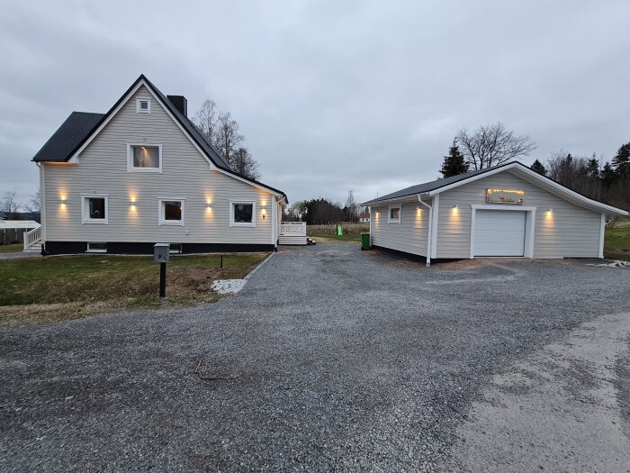 Ett grått trähus med svarta detaljer och fönsterljus. Bredvid finns en fristående garagebyggnad, omgiven av natur. Grusväg leder till egendomen.