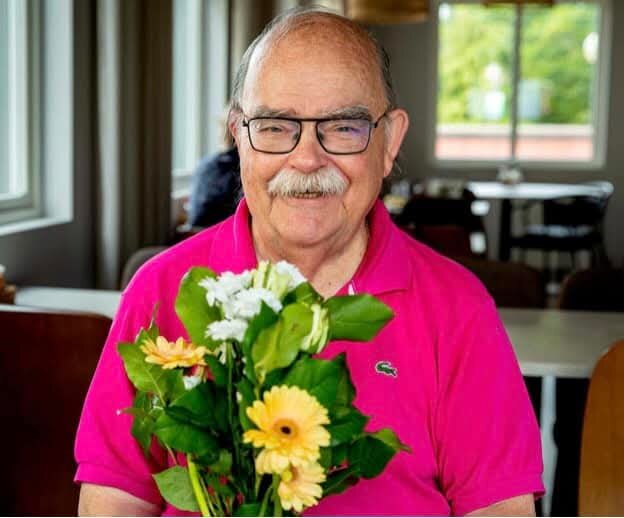 En äldre man i rosa tröja håller en blombukett med gula och vita blommor, sitter inomhus vid ett bord.