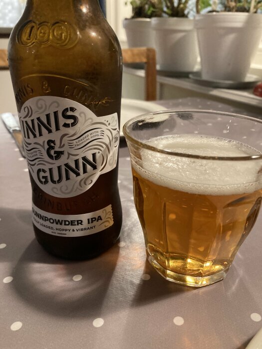 Ölflaska och glas med skummande IPA på prickigt bord, växter i bakgrunden.