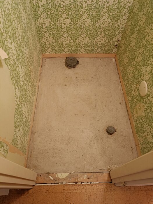 En renslipad betonggolvyta i en liten toalett, omgiven av grönblomstrande tapet på väggarna.