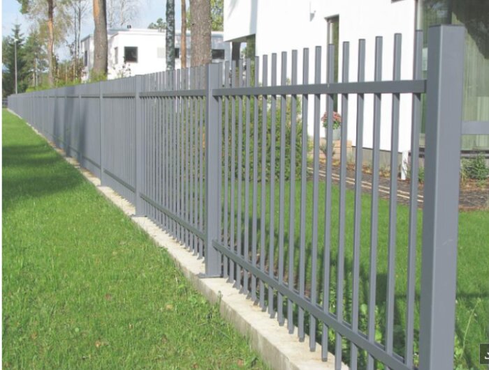 Svart smidesstaket ovanpå en låg mur av betongsten, längs en grönskande trädgård, med modern byggnad i bakgrunden.