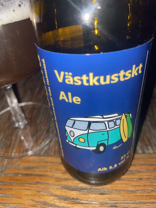 Flaska med blå etikett märkt "Västkustskt Ale", placerad bredvid ett glas med mörk öl.