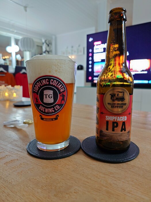 Ett glas öl med etiketten "Toppling Goliath" och en ölflaska märkt "Shipfaced IPA" på ett bord i ett modernt vardagsrum.