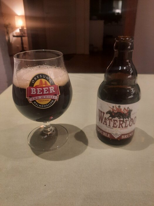 Ett glas mörk öl med texten "Oresund Beer and Whisky Festival" bredvid en flaska märkt "Waterloo Double Brun" på ett bord.