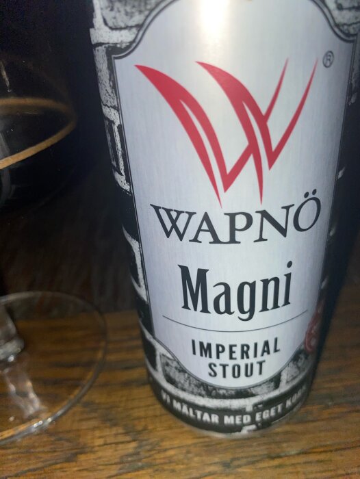 Burk av Wapnö Magni Imperial Stout öl, placerad bredvid ett glas på ett träbord.